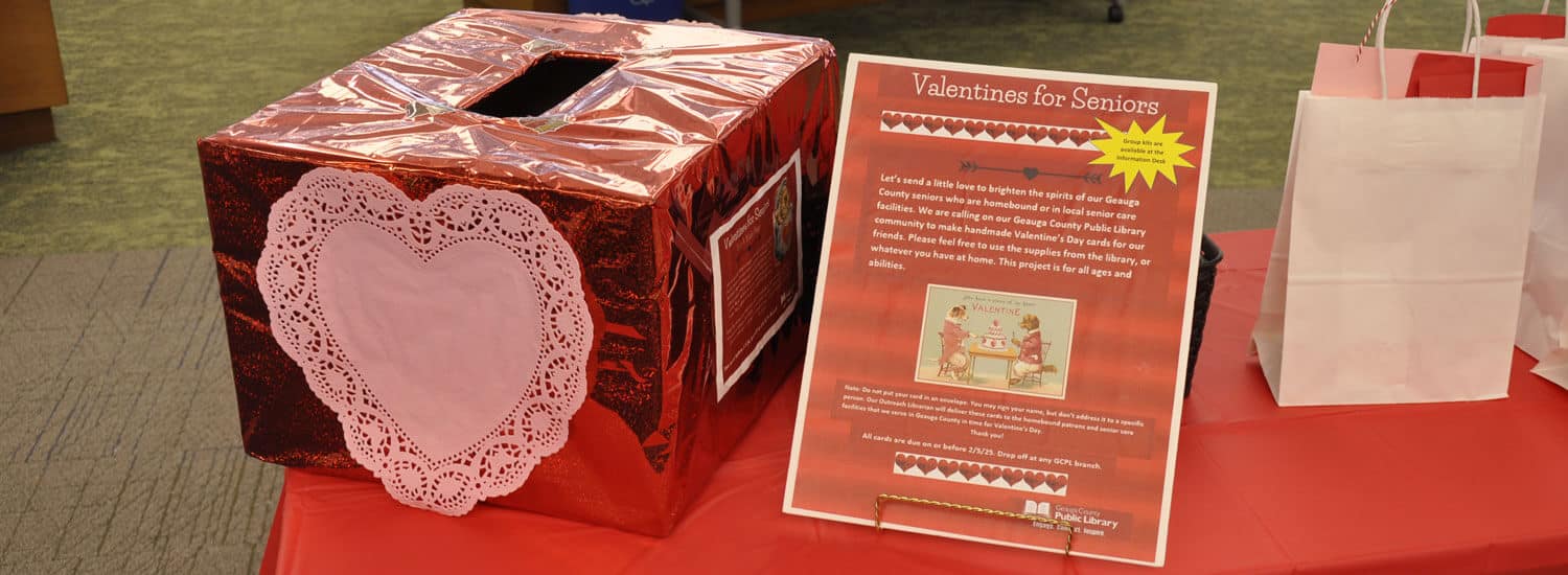A "Valentines for Seniors" poster and submission box sit on a table draped with red cloth.