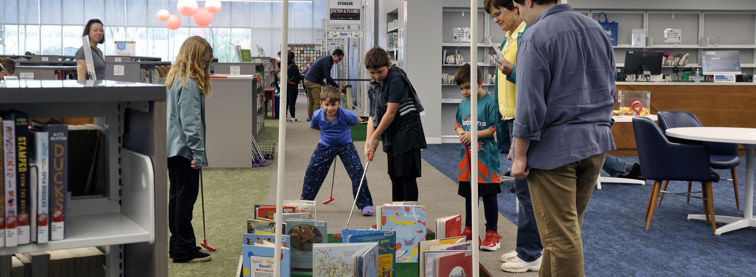Mini-Golf at the Library, Family Event A young boy plays mini-golf at Geauga County Public Library’s Bainbridge Branch while a group of children and adults watch nearby.